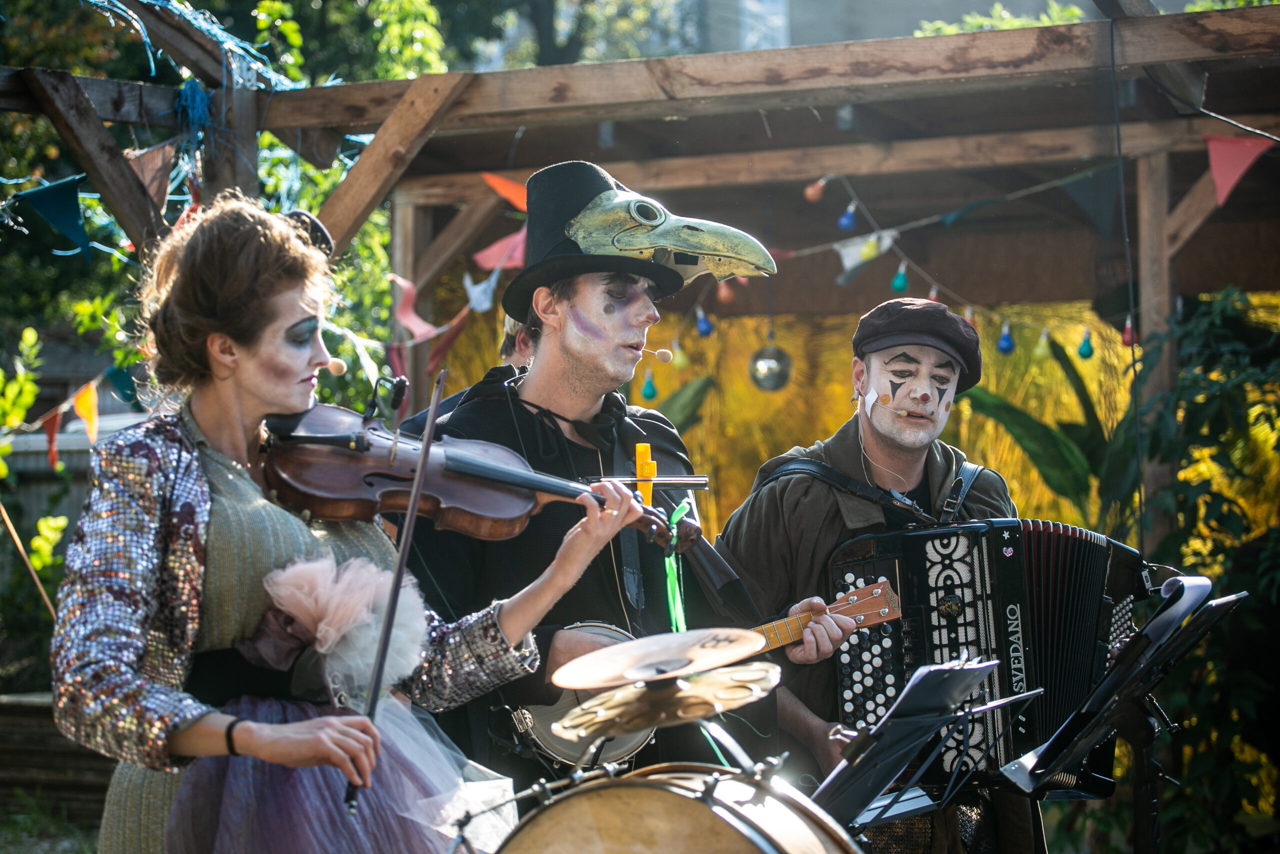 Zdjęcie. Od lewej stoją skrzypaczka, muzyk grający na banjo i akordeonista ubrani w fantazyjne stroje i mający pomalowane twarze. Występują na świeżym powietrzu. W tle drewniana konstrukcja altany, rośliny i kolorowe lampki.